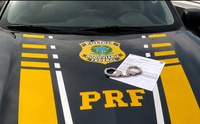 Imagem institucional da Polícia Rodoviária Federal (PRF). Sobre o capô de uma viatura oficial da PRF, identificado com faixas amarelas, brasão institucional e a sigla “PRF”, há um documento impresso parcialmente coberto por um par de algemas metálicas. A fotografia foi registrada em ambiente externo e não apresenta pessoas visíveis.