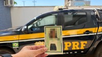 Fotografia colorida feita em ambiente externo e iluminado pela luz do dia. No centro inferior da imagem, uma pessoa segura um documento de identidade brasileiro com uma das mãos. O documento está próximo à câmera e ocupa o foco principal da cena; a fotografia e as informações pessoais aparecem desfocadas para preservar a privacidade. Ao fundo, ocupando grande parte da imagem, há uma viatura da Polícia Rodoviária Federal nas cores preta e amarela. A sigla “PRF” está visível na porta traseira do veículo. Também é possível ver uma pequena bandeira do Brasil na lateral. O fundo apresenta um muro claro, parte de uma construção e céu azul. A profundidade de campo reduzida mantém o documento em destaque enquanto o veículo e o ambiente aparecem desfocados.
