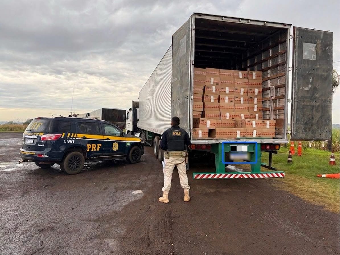 A imagem mostra um policial e uma viatura ao lado de um caminhão carregado com caixas de cigarro