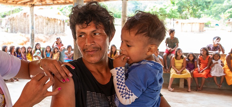 Indígena sendo vacinado com uma criança no colo