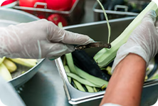 Mãos com luvas descascando vegetais em uma cozinha industrial.