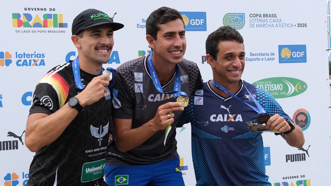 Caio Bonfim e mais dois atletas masculinos sorriem no pódio da Copa Brasil de Marcha Atlética 2026. Os atletas seguram suas medalhas e, ao fundo, um painel apresenta as marcas de patrocinadores e entidades esportivas.