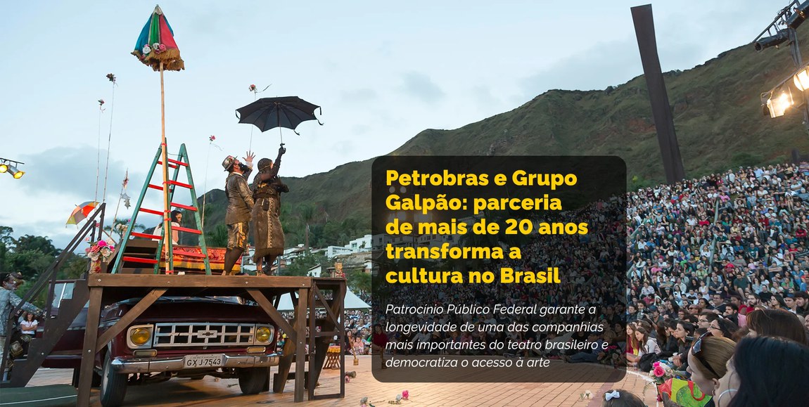 Apresentação teatral ao ar livre com grande público sentado em uma encosta. O palco é montado sobre um caminhão Chevrolet antigo, decorado com guarda-chuvas coloridos, escada e adereços vibrantes. Dois atores em figurinos elaborados estão em cena, um deles segurando um guarda-chuva. Ao fundo, vê-se montanhas e construções urbanas. O espetáculo é parte da parceria entre Petrobras e o Grupo Galpão, que há mais de 20 anos promove a democratização do acesso à arte e fortalece a cultura brasileira por meio de patrocínio público federal.