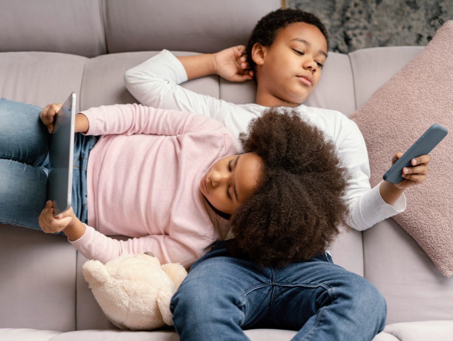 Imagem quadrada com duas crianças usando um tablet, sendo um garoto preto, com cabelo curto e preto, segurando o aparelho, e uma garota branca, com cabelos longos e castanhos, observando, ambos sorrindo e com blusas em tons claros