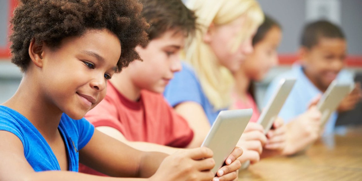 Grupo de crianças em uma sala de aula usando tablets. Em destaque, uma menina de cabelo encaracolado e camiseta azul sorri levemente enquanto segura o dispositivo, ao lado de outras crianças concentradas em seus tablets. A cena sugere um ambiente de aprendizado interativo e digital