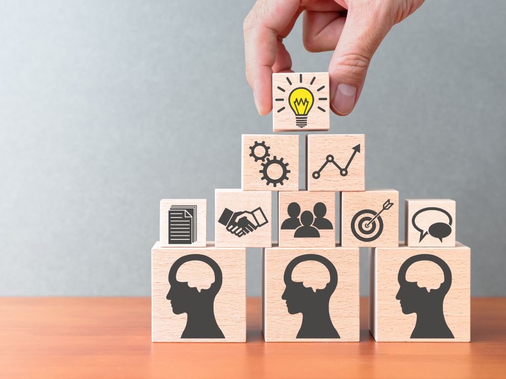 A imagem mostra um conjunto de blocos de madeira dispostos em forma de pirâmide sobre uma mesa, com um fundo cinza. Na base, há cinco blocos, cada um com a silhueta de uma cabeça humana de perfil.