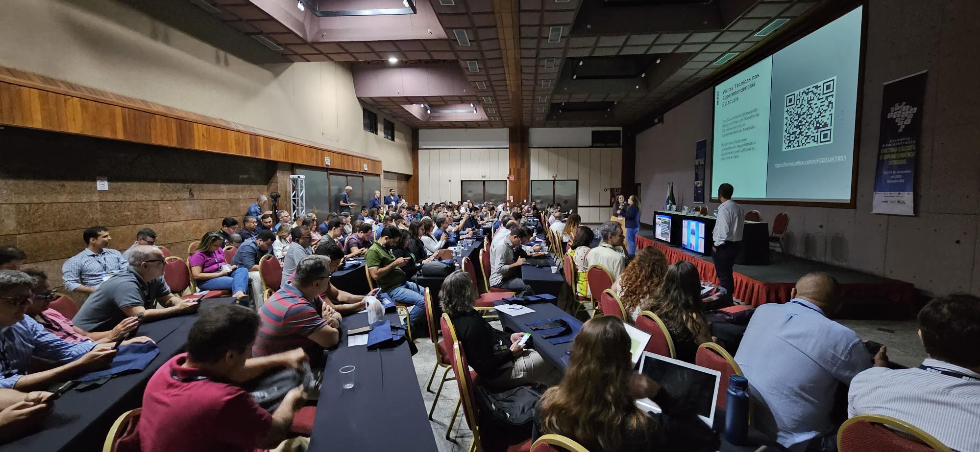 Grande auditório de teto escuro quadriculado e detalhes de madeira nas paredes, com várias fileiras de mesas longas azul-escuras alinhadas de lado a lado nesse espaço. Cada mesa tem três ou quatro cadeiras vermelhas, e quase todas estão ocupadas pelo público. À direita da foto, há um palco com uma grande tela de projeções, em frente ao qual três pessoas estão de pé; uma delas, uma mulher, está falando ao microfone olhando para a platéia.