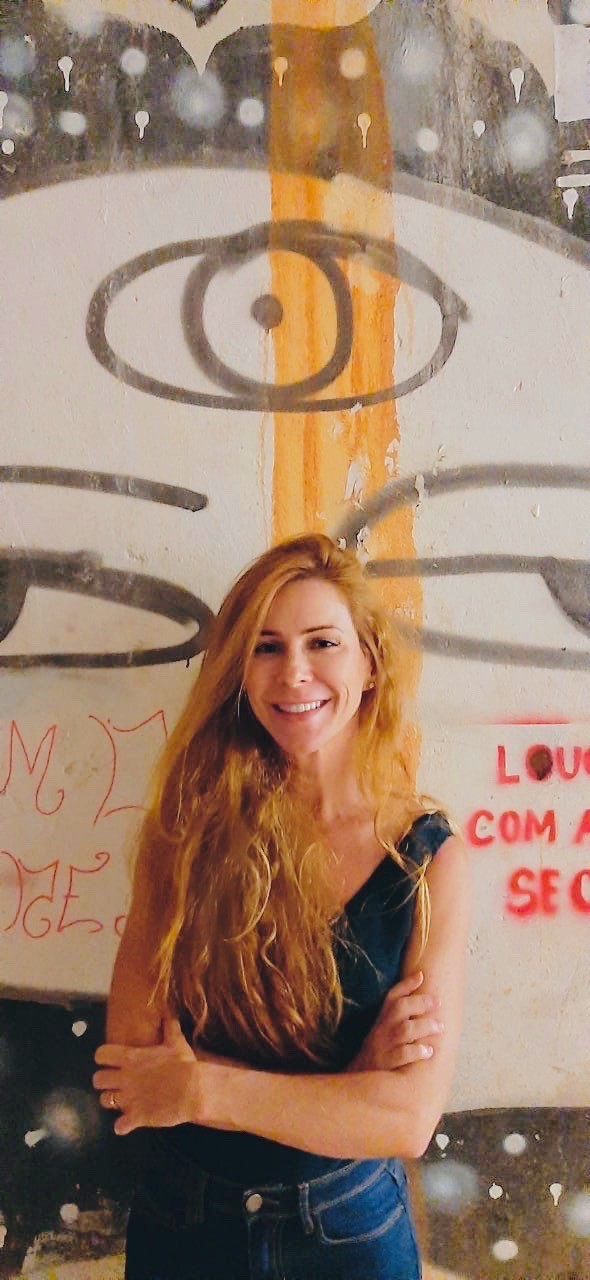 mulher branca, cabelos lisos, longos e ruivos. Sorri para a foto e veste camiseta preta e jeans. Fundo uma parede grafitada