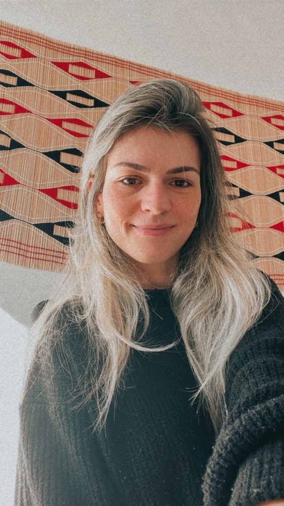 mulher branca, cabelos lisos, loiros e longos. Sorri para a foto e veste um casaco de lã cinza. Em seu fundo, uma parede branca ornada com um quadro com losangos coloridos.