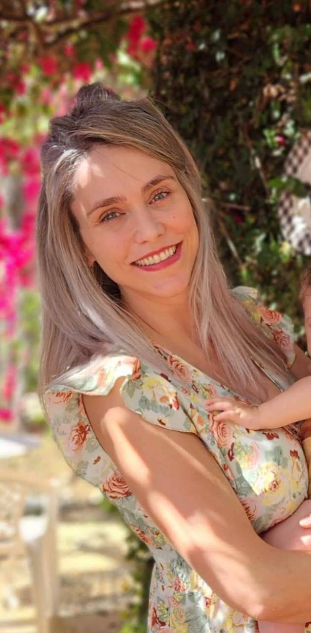 mulher branca, olhos azuis, cabelos lisos, loiros, abaixo dos ombros, trajando um vestido sem manga amarelo claro, sorri para a foto. Ao fundo, uma árvore com flores.