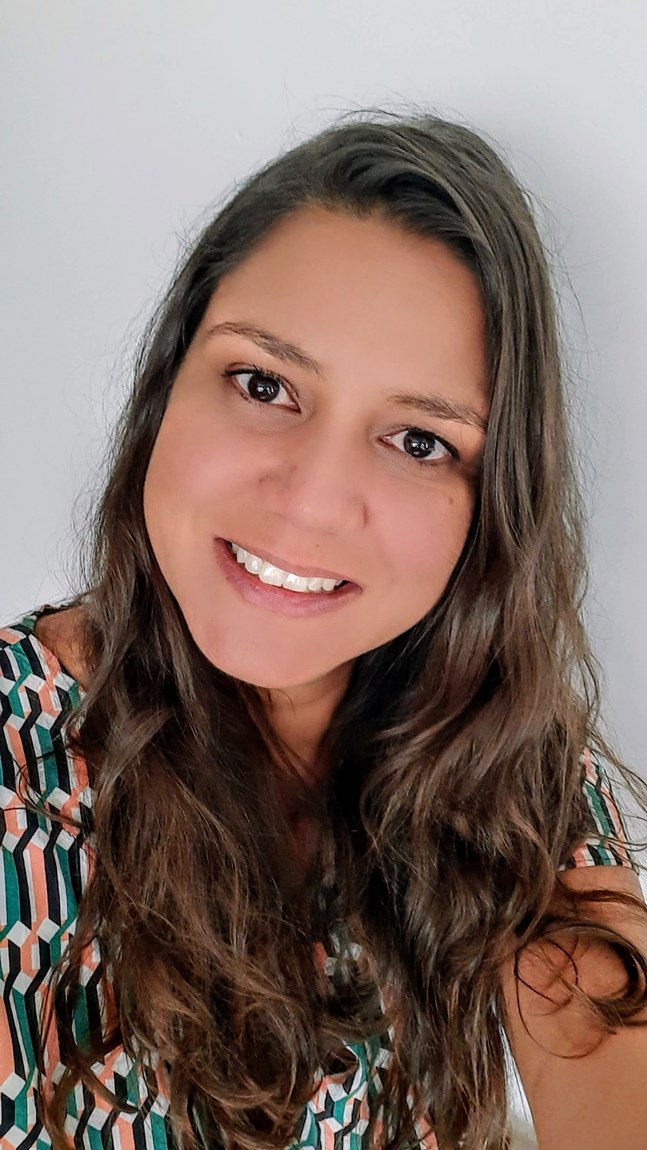 Mulher parda, olhos pretos, cabelos castanhos ondulados abaixo dos ombros, sorriso leve no rosto, vestindo uma blusa estampada com figuras geométricas verdes, laranjas, brancas e pretas; fundo branco