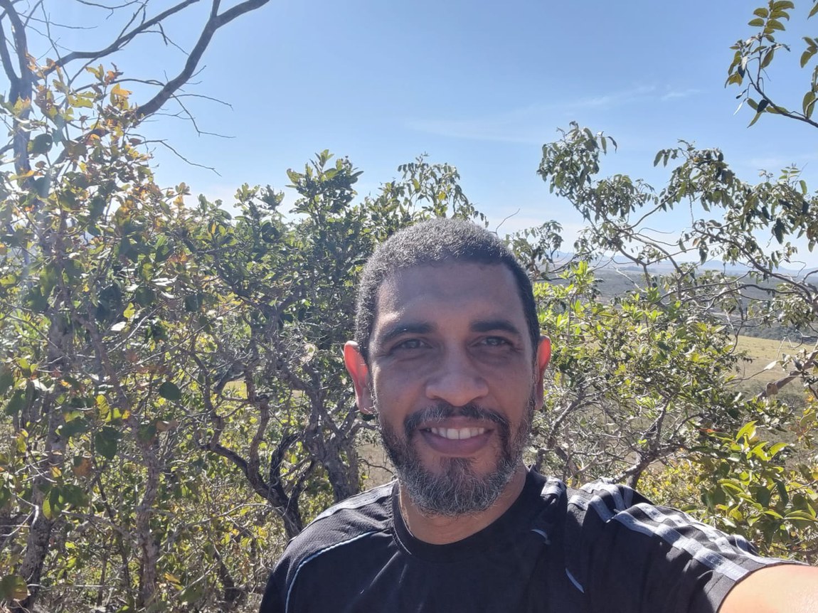 homem preto, cabelos curtos, barba curta, trajando camiseta preta sorri para a câmera em meio à natureza