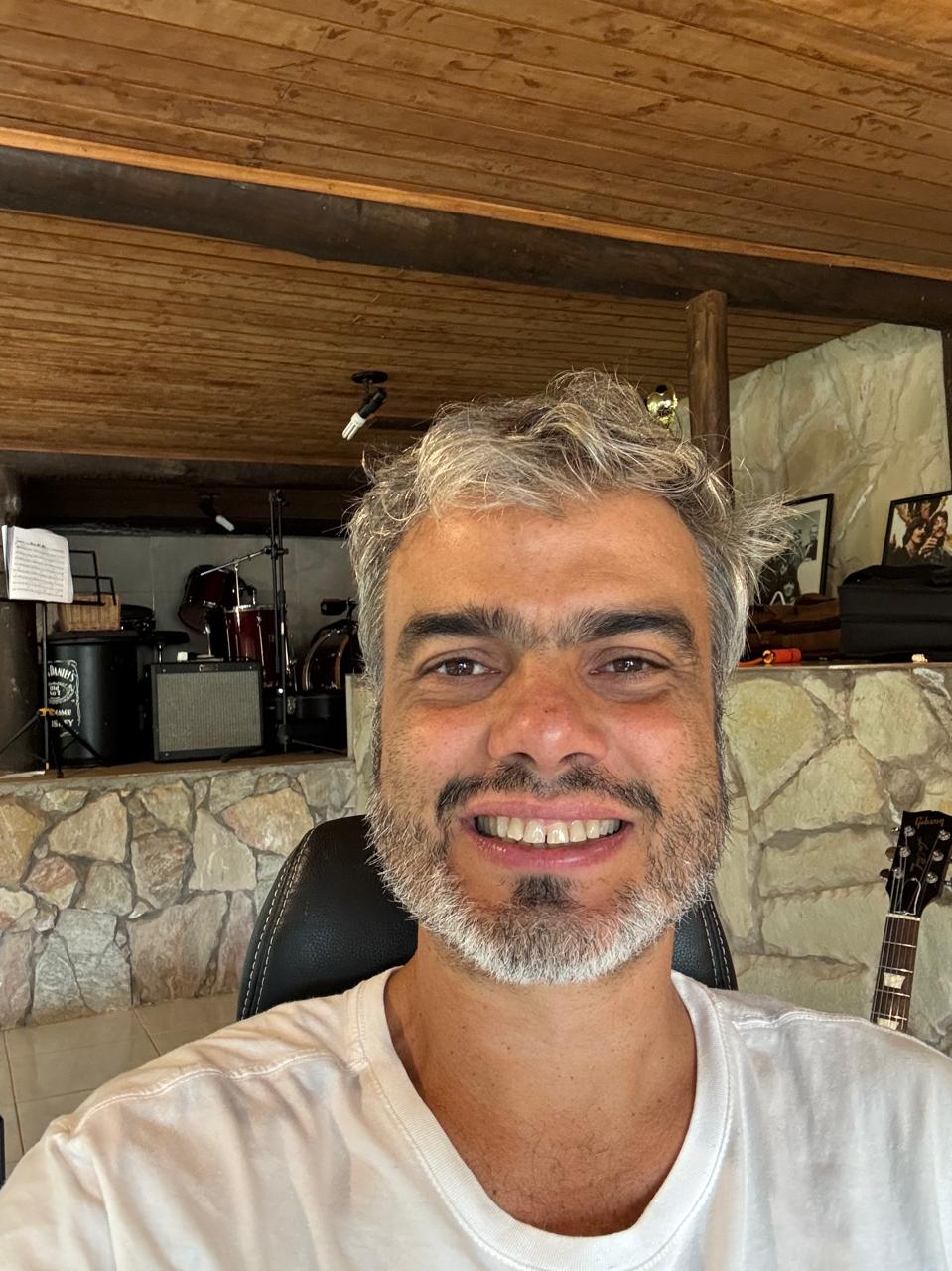 Homem branco de cabelos curtos e grisalhos, sorri com barba também grisalha, veste uma camiseta branca. Atrás um fundo de pedra e teto de madeira com alguns objetos