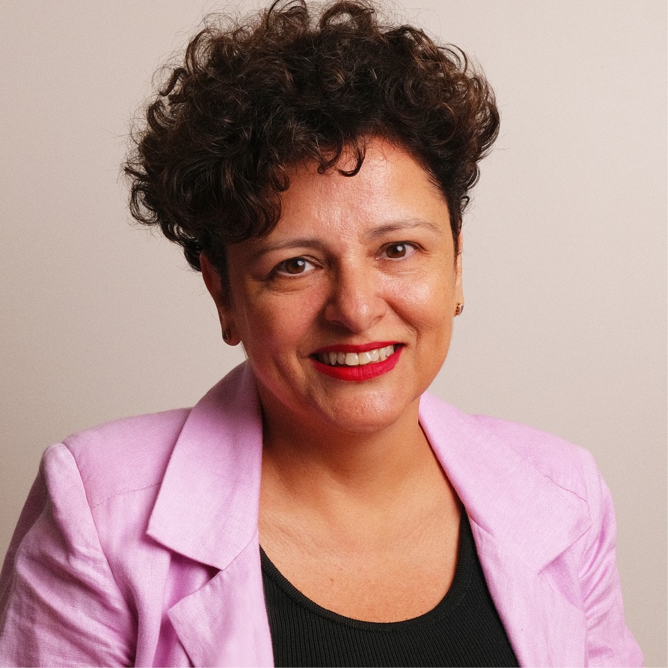 Joseane, mulher branca com cabelo curto e cacheado, usando blazer rosa e blusa preta, sorrindo para a câmera, em fundo claro e neutro.”