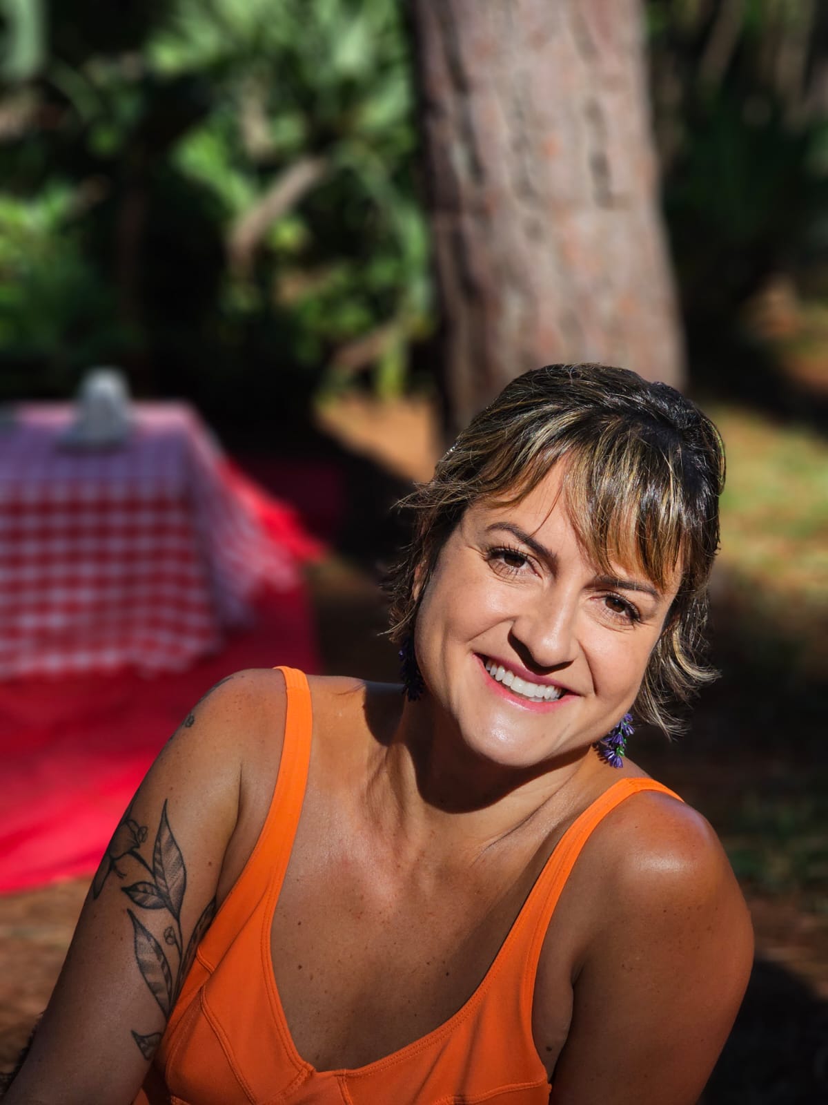 mulher branca de cabelos curtos e castanhos vestindo uma blusa de alça laranja. Está sentada e sorrindo. Ao seu fundo um gramado, uma árvore e uma mesa baixa com toalha quadriculada em vermelho e branco sobre um tapete vermelho.