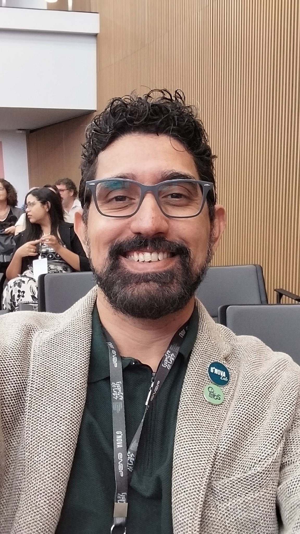 homem pardo de óculos, ovais, cabelos pretos curtos, barba, trajando blazer cáqui e camisa verde escura.
