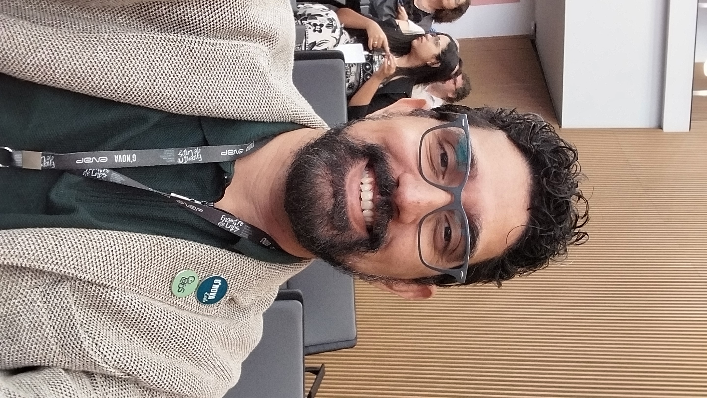 homem pardo de óculos, ovais, cabelos pretos curtos, barba, trajando blazer cáqui e camisa verde escura.