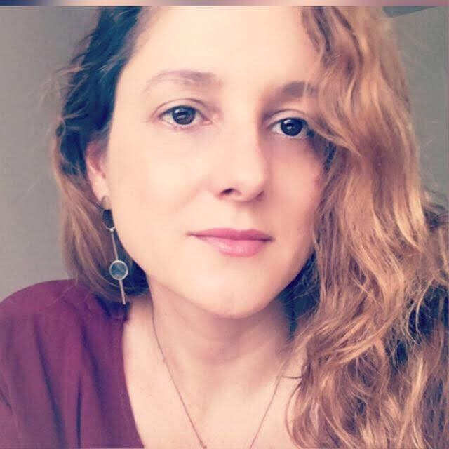 Mulher branca com cabelos ruivos e compridos e olhos castanhos. Está vestido uma blusa roxa e sorri levemente.