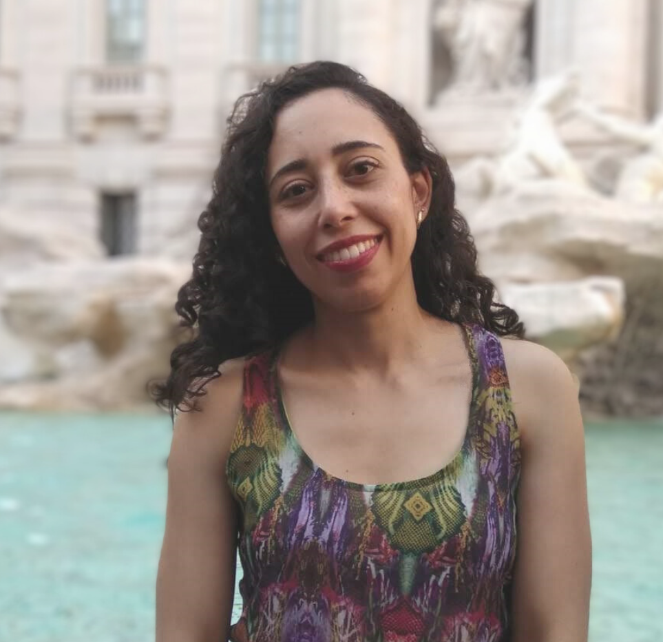 mulher branca, cabelos cacheados pretos e longos. Sorri para a foto e veste uma blusa de alça colorida. Em seu fundo esculturas brancas e uma fonte.