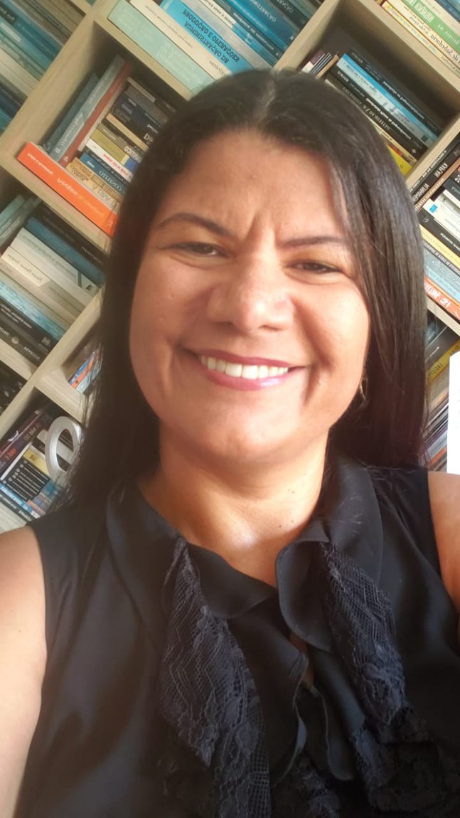 Mulher parda, cabelos alisados castanhos escuros repartidos ao meio, cobrindo as orelhas e na altura dos ombros. Rosto ovalado com a testa franzida ao centro. Olhos castanhos escuros, sombrancelhas arqueadas, nariz largo e sorriso aberto. Veste blusa preta, com babado na gola deixando os ombros à mostra. Ao fundo uma estante de livros.