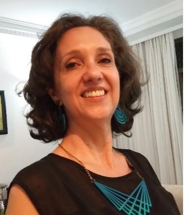 Mulher branca, cabelos castanhos ondulados na altura dos ombros e sorriso no rosto. Ao fundo uma sala de estar com persiana branca