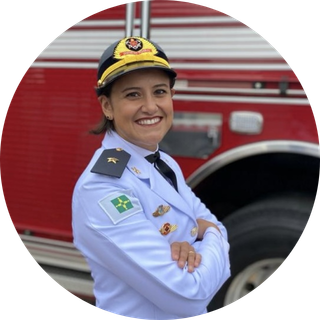 Mulher usando uniforme branco do Corpo de Bombeiros e boné com brasão da corporação. Ela está de braços cruzados, sorrindo, em frente a um caminhão vermelho dos bombeiros.