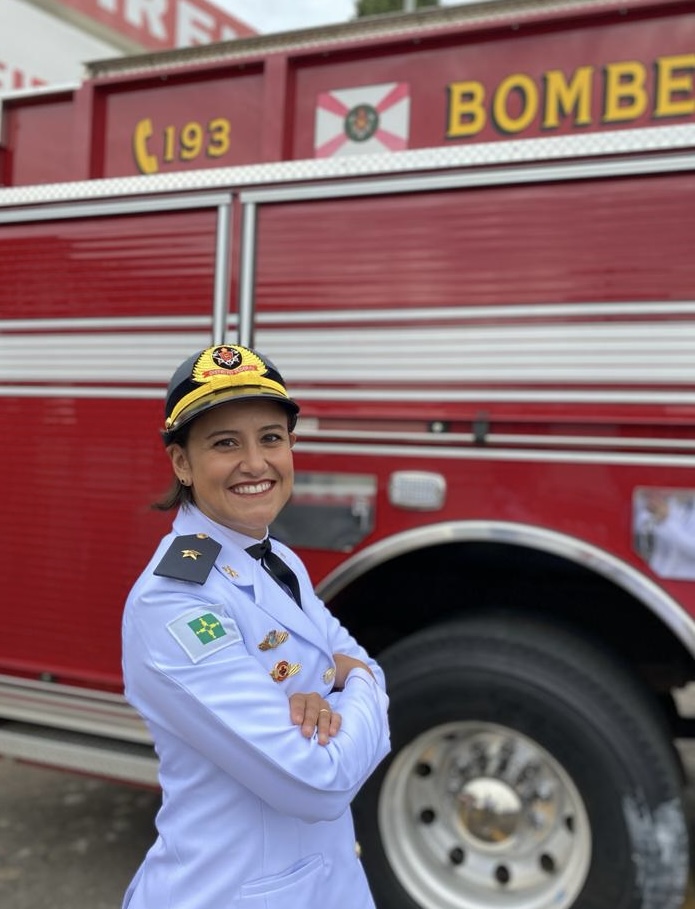 Mulher usando uniforme branco do Corpo de Bombeiros e boné com brasão da corporação. Ela está de braços cruzados, sorrindo, em frente a um caminhão vermelho dos bombeiros.
