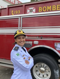 Mulher usando uniforme branco do Corpo de Bombeiros e boné com brasão da corporação. Ela está de braços cruzados, sorrindo, em frente a um caminhão vermelho dos bombeiros.