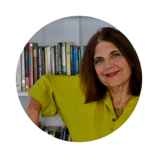 mulher branca, cabelos castanhos até o ombro. Ela está em pé, sorrindo, usando uma blusa verde e apoiada em uma estante cheia de livros