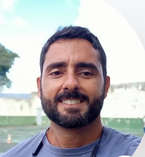 Homem com barba curta e cabelo escuro, sorrindo levemente. Ele veste uma camiseta cinza e está ao ar livre, com um muro e árvores ao fundo.