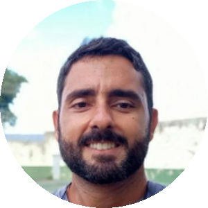 Homem com barba curta e cabelo escuro, sorrindo levemente. Ele veste uma camiseta cinza e está ao ar livre, com um muro e árvores ao fundo.