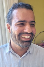 foto do rosto de perfil de Rafael Xucuru-Kariri. Um homem indígena, de cabelos e olhos escuros e barba baixa. Ele veste uma camisa de botão cinza clara. Em seu fundo uma parede amarela e uma cortina branca.