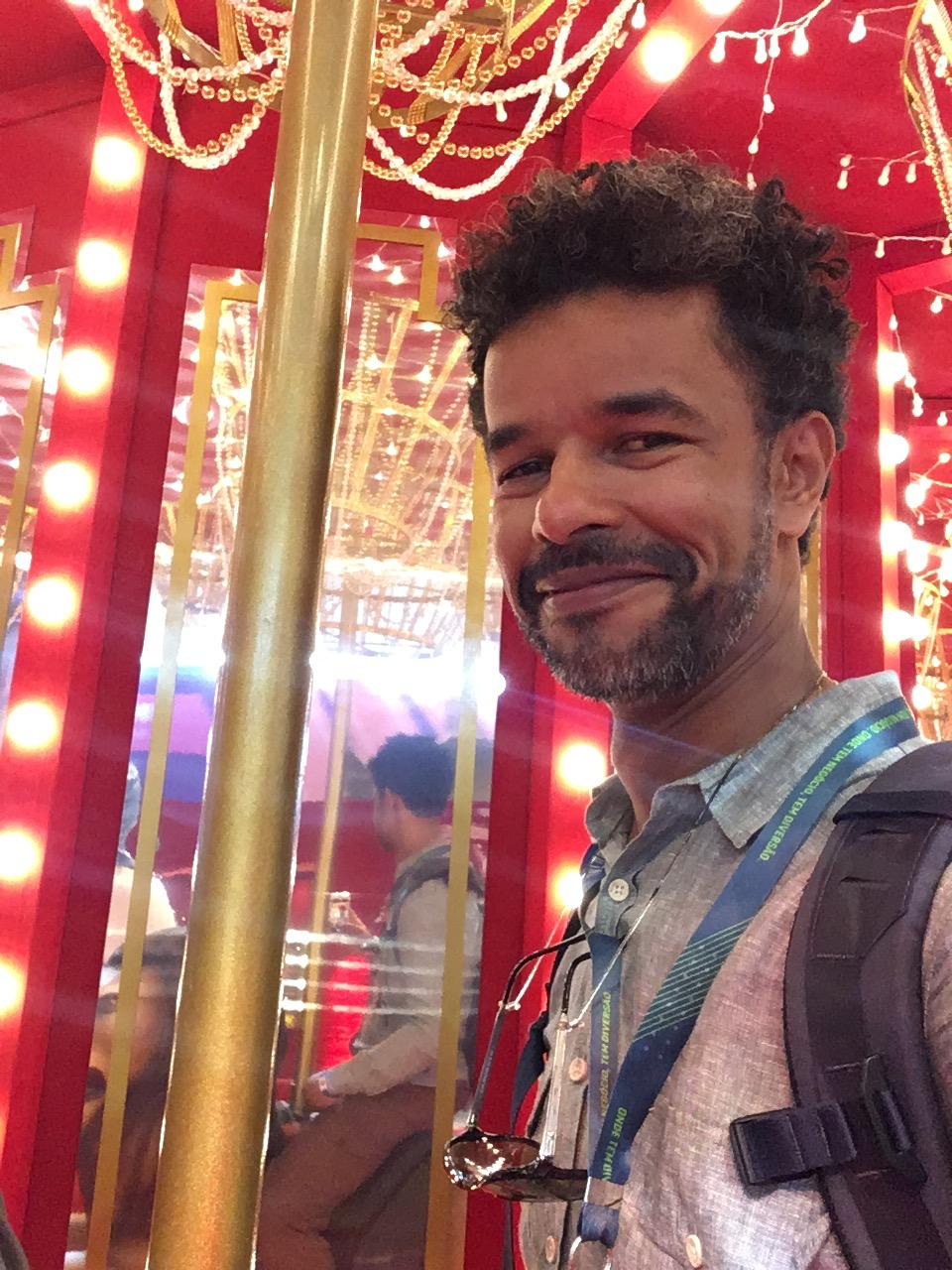 homem pardo, olhos castanho-escuros, barba, cabelo raspado nas laterais e cachos pretos no topo. ele veste uma camisa branca com uma mochila preta. tem um óculos de sol e um crachá pendurado no pescoço. sorri levemente. está em uma sala vermelha espalhada.