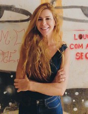 Mulher branca de cabelos comprido, ruivo e ondulado. Olhos castanhos. Vestindo uma blusa sem manga, preta, e calça jeans. Está em pé, de braços cruzados e sorrindo. Ao seu fundo, uma parede com grafite, em uma passagem subterrâneas sob o Eixão, em Brasília.