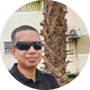 Fotografia vertical de Odenilton Júnior, da cintura para cima. É um homem negro, de pele marrom escura e cabelo preto bem curto. De rosto ovalado, usa óculos escuros retangulares, tem nariz largo e lábios grossos. Sorri discretamente. Usa camiseta polo preta de mangas curtas, com detalhe em cinza nas mangas e nos ombros, onde se lê "Calvin Klein". Os braços cruzados à frente do corpo têm uma pulseira prateada no pulso esquerdo.