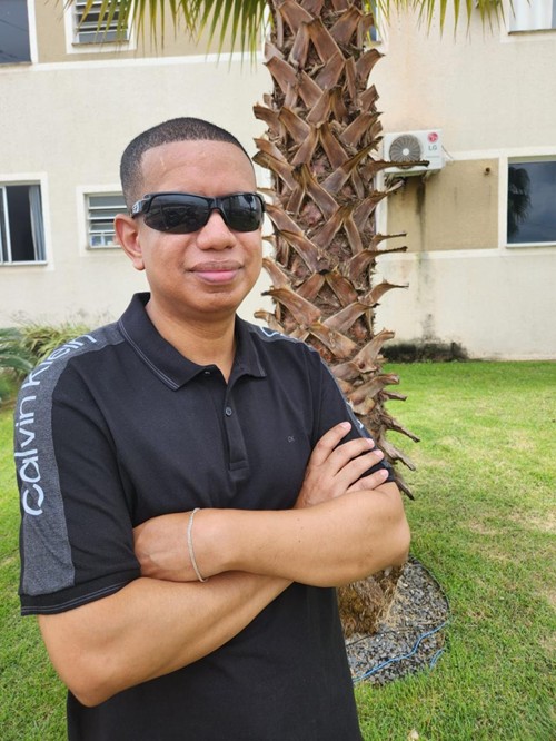 Fotografia vertical de Odenilton Júnior, da cintura para cima. É um homem negro, de pele marrom escura e cabelo preto bem curto. De rosto ovalado, usa óculos escuros retangulares, tem nariz largo e lábios grossos. Sorri discretamente. Usa camiseta polo preta de mangas curtas, com detalhe em cinza nas mangas e nos ombros, onde se lê "Calvin Klein". Os braços cruzados à frente do corpo têm uma pulseira prateada no pulso esquerdo.