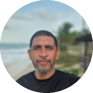 Marcelo Ivo, homem negro, de pele parda, olhos castanhos e cabelos crespos, escuros e curtos. Ele tem uma barba grisalha e curta, usa uma camisa preta e ao fundo vê-se o mar.