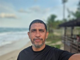 Marcelo Ivo, homem negro, de pele parda, olhos castanhos e cabelos crespos, escuros e curtos. Ele tem uma barba grisalha e curta, usa uma camisa preta e ao fundo vê-se o mar.