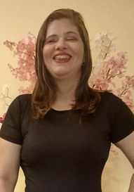 Doriane Vasconcelos, uma mulher branca, de cabelos claros, na altura dos ombros e olhos semifechados. Doriane veste uma blusa preta, usa batom vermelho e sorri. Ao fundo, um papel de parede bege com flores rosas.