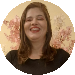 Doriane Vasconcelos, uma mulher branca, de cabelos claros, na altura dos ombros e olhos semifechados. Doriane veste uma blusa preta, usa batom vermelho e sorri. Ao fundo, um papel de parede bege com flores rosas.