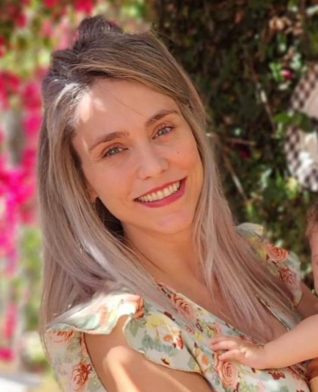 Isabela Blumm, uma mulher branca, de olhos azuis, cabelos lisos e loiros. Ela está em pé, sorrindo e usa um vestido estampado florido de cores claras.