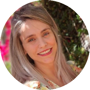 Isabela Blumm, uma mulher branca, de olhos azuis, cabelos lisos e loiros. Ela está em pé, sorrindo e usa um vestido estampado florido de cores claras.