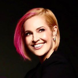 Mulher branca, cabelos curtos e lisos na cor loira com mexa rosas. Ela sorri olhando para o horizonte.