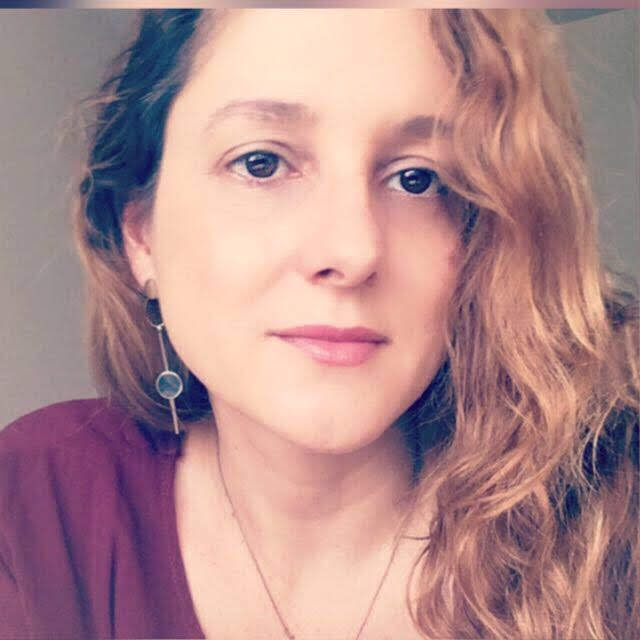 Mulher branca com cabelos ruivos e compridos e olhos castanhos. Está vestido uma blusa roxa e sorri levemente.