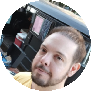 Homem com barba curta e cabelo castanho claro, vestindo uma camiseta amarela com estampa. Ele sorri levemente e está em frente a uma kombi preta com objetos no interior.