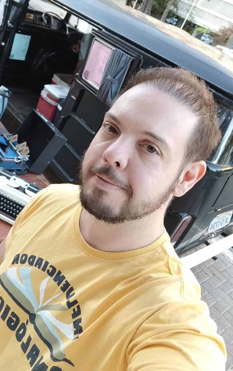 Homem com barba curta e cabelo castanho claro, vestindo uma camiseta amarela com estampa. Ele sorri levemente e está em frente a uma kombi preta com objetos no interior.