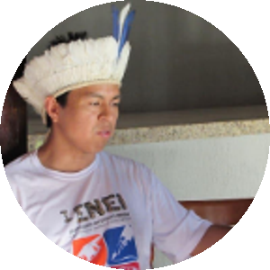 rosto de perfil de Jibran Yopopem Patté, um homem indígena, de olhos e cabelos escuros. Ele veste uma camiseta branca e usa um cocar com penas brancas e uma pena azul ao centro.