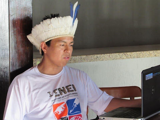 rosto de perfil de Jibran Yopopem Patté, um homem indígena, de olhos e cabelos escuros. Ele veste uma camiseta branca e usa um cocar com penas brancas e uma pena azul ao centro.