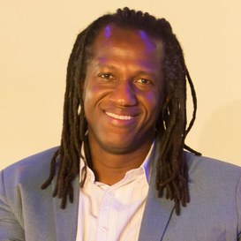 homem preto sorrindo, olhos pretos e cabelos trançados até o ombro. Veste um terno cinza e uma camisa branca.