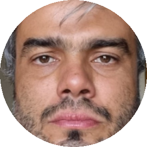 Homem branco de cabelos curtos e grisalhos, sorri com barba também grisalha, veste uma camiseta branca. Atrás um fundo preto.